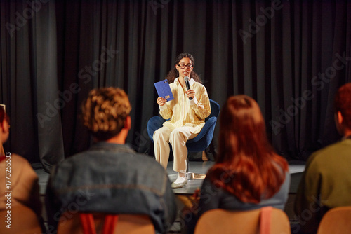 Young adult woman sitting on stage holding microphone and book, speaking to audience of young adults, engaging in public presentation or discussion event