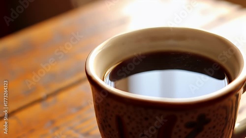 Close up of a warm mug filled with dark aromatic coffee sitting on a rustic wooden table bathed in soft morning sunlight with subtle reflections on the surface of the beverage