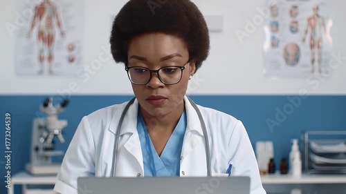 Black Female Doctor Wearing Glasses And Stethoscope Concentrating On Laptop Screen In A Medical Office With Anatomical Charts On Wall
