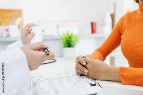 A doctor is showing a patient a bottle of medicine. The patient is holding a pen and writing something down. The doctor is wearing a white coat and the patient is wearing an orange sweater
