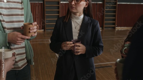 Tilt down of young female psychologist and teenagers with cups of tea or coffee discussing main points of forthcoming session while standing in circle in sports hall