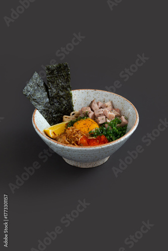 A bowl of Mazesoba topped with pork, egg yolk, green onions, Nori, flying fish and lemon on black background.