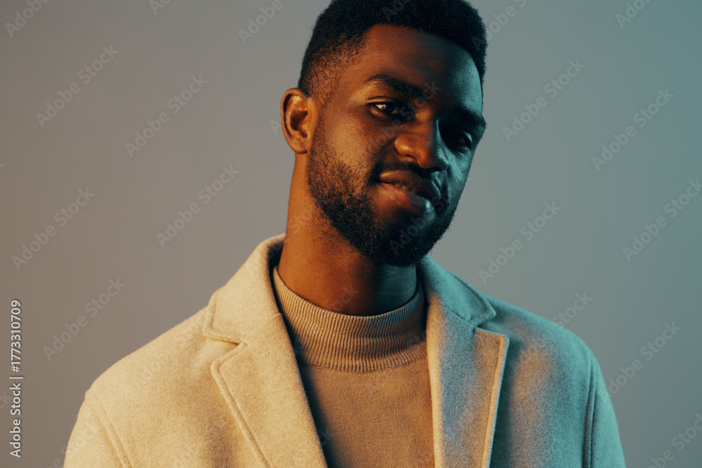 Obraz premium Stylish young man with a confident expression, wearing a light coat against a contrasting blue and beige background, showcasing contemporary fashion and personal style