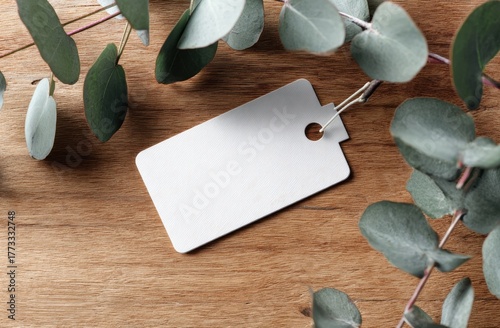 Blank white tag with hole, surrounded by eucalyptus leaves on wooden