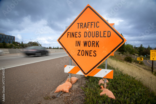 Orange traffic sign stating Traffic Fines Doubled in Work Zones with traffic in the background