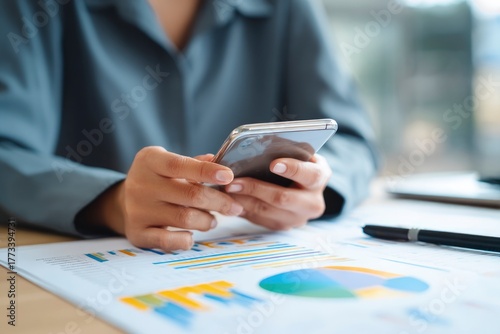 A focused close-up of hands interacting with a sleek smartphone, positioned above a desk adorned with colorful business charts and financial reports. This image effectively embodies modern digital com