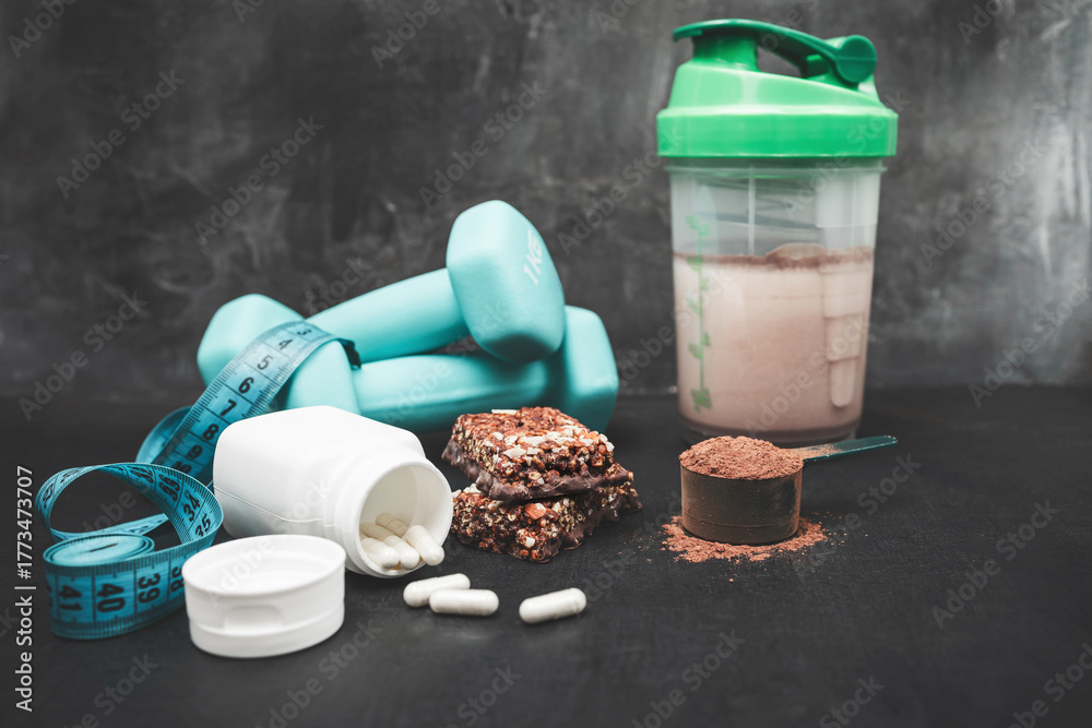 Naklejka premium Chocolate whey protein powder scoop, white amino acid capsules, protein shake in shaker bottle, protein bar, dumbbells, measuring tape on dark background, sport nutrition and healthy lifestyle concept