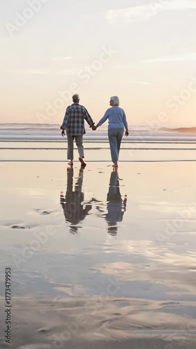  two individuals walking on beach at sunset. person in foreground is wearing plaid shirt and light-colored pants, while individual behind