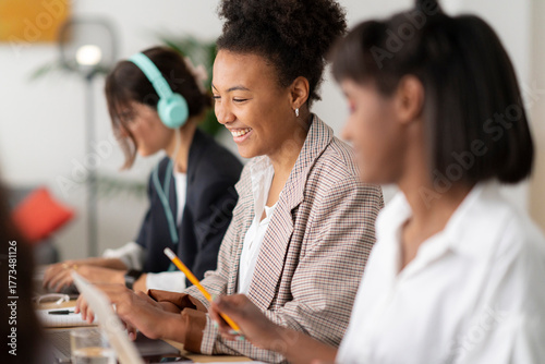 Diverse young women are working together in a modern office setting. The central woman smiles brightly, holding a pencil and looking at her laptop. Another woman wears headphones, focused on her task.