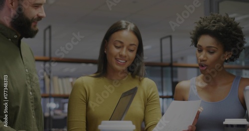 Three colleagues collaborating at table in office, with bookshelves, tablet, papers and coffee cups