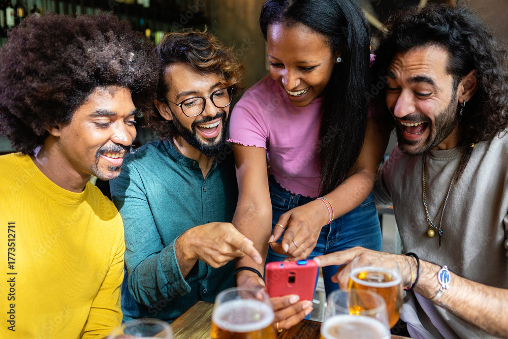 Obraz premium Cheerful multi-ethnic friends having fun together, drinking beer and using a smartphone at the bar