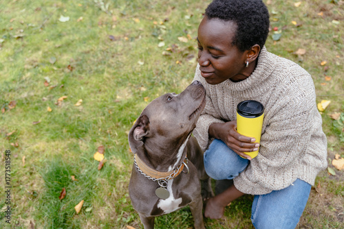 Black woman with pit bull dog and yellow cup sitting on grass outdoors in autumn park