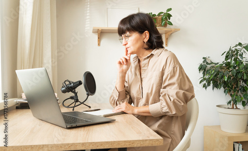 Mature caucasian woman podcasting from home office with laptop and microphone setup