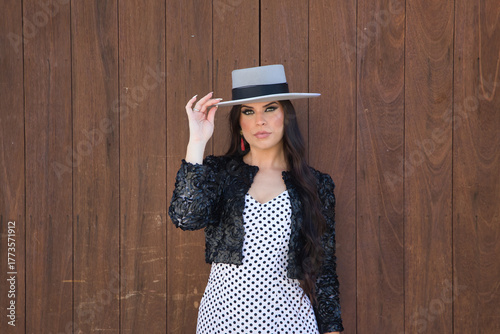 Portrait of young woman, beautiful, brunette, flamenco dancer, wearing beautiful dress with jacket and hat, posing with wooden door in background. Flamenco concept, dancer, typical, Spanish, Seville.