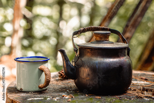 An old, dark metal kettle and a white enamel mug sit on a log table outdoors. The photo has a soft, warm, earthy tone with a pleasant green bokeh background, showing camp life