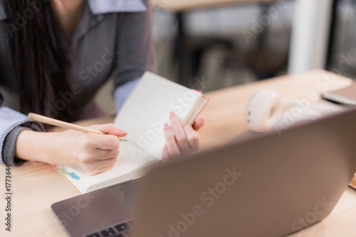 Woman's hands writing notes in an open notebook with a pencil, focusing on tasks while using a laptop and headphones on a desk, representing online learning and remote work concepts
