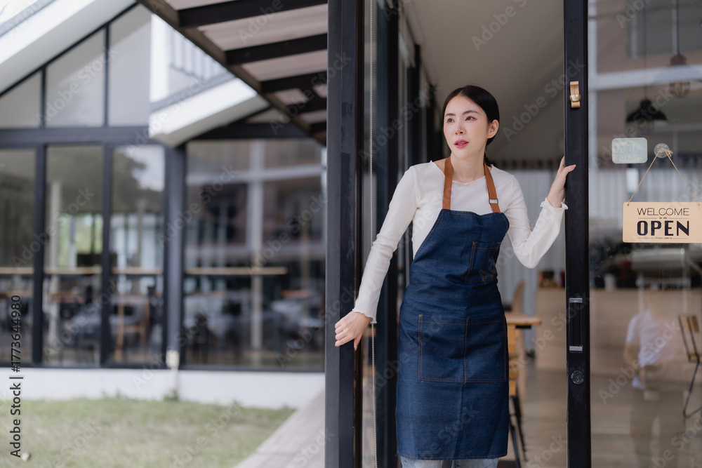 Fototapeta premium Young Asian woman small business owner wearing an apron, opening the glass door of her coffee shop with an open sign, ready to welcome customers for the day