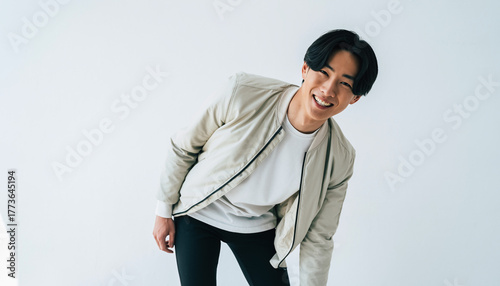 純白のスタジオ背景に立つ20代の日本人男性のポートレート。白いTシャツにライトベージュのボンバージャケットを重ね、黒のスリムパンツで引き締めたシンプルで都会的なスタイル。短い黒髪と明るくエネルギッシュな笑顔、少し前傾した躍動感ある姿勢が、爽やかで清潔感のあるムードを演出。