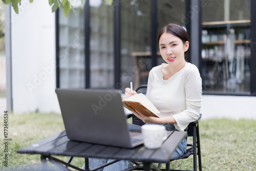 Asian woman smiling while remote working outdoors, sitting by a laptop and writing in a notebook, demonstrating flexibility and modern work-life balance in a home garden setting