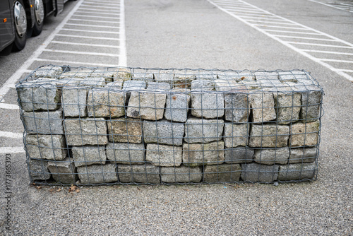 A gabion wall made of gray cut stones stacked inside a rectangular wire mesh cage on the parking lot floor. Landscape architecture.