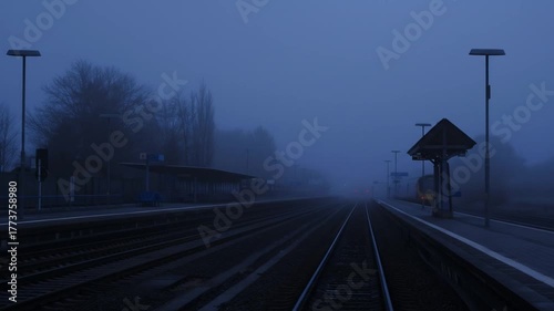 Empty train station shrouded in dense blue fog at dawn, with multiple railway tracks and platform disappearing into the mysterious mist, eerie atmosphere.