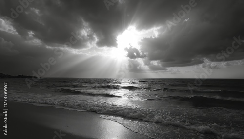 Dramatic sun rays pierce dark clouds over ocean waves hitting sandy shore. Black and white coastal scene evokes moody atmosphere. Horizon line divides sky and sea. Peaceful twilight view.