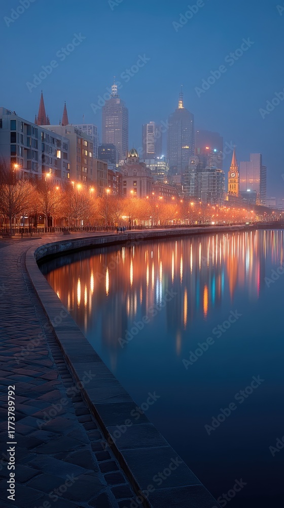 Naklejka premium Cityscape at night with illuminated skyline reflected in calm water during dusk with warm streetlights and buildings lining the riverbank with a clear blue and orange sky.