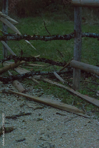 A wooden split-rail fence with broken and moss-covered sections borders a gravel path next to a grassy area and dark trees in the evening