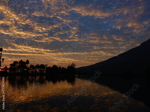 The dawn atmosphere with golden rays of sun in the blue sky, silhouettes of trees, and reflections on the lake water create a calming combination of views.