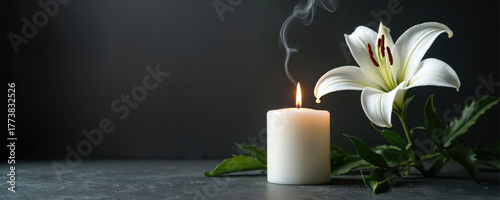 White lily flower and burning candle on dark surface. Gentle smoke rises from candle flame. Space for text offers solemn remembrance or condolence message for loss.