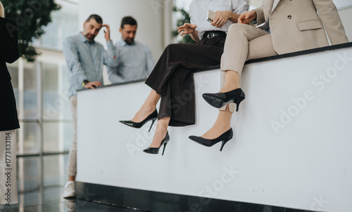 A group of professionals relaxes on a white ledge in a bright office lobby, dangling their legs in stylish heels. They appear focused yet at ease, pausing between meetings.