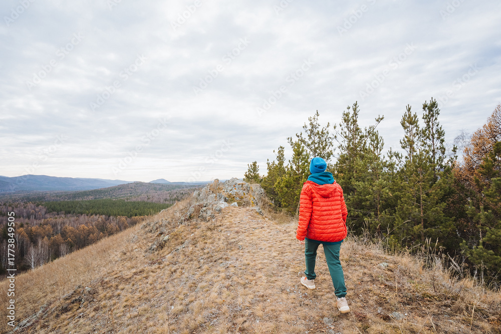Naklejka premium Young man on mountain trail, Young person walks along solitary mountain ridge at dusk, Caucasian teenage boy carefully navigates narrow ridge through peaceful valley scenery with overcast