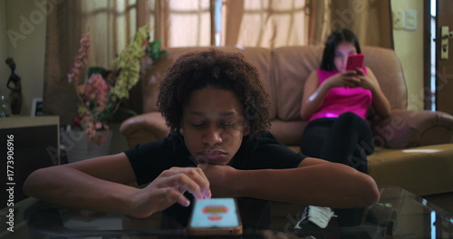 Young Boy with curly hair lying on glass table scrolling smartphone, disengaged, while another young person sits on sofa in background on phone, themes of social media addiction and isolation