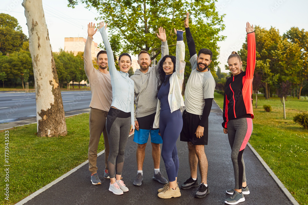 Fototapeta premium Happy sporty young people in sportswear standing with hands up after successful sport exercises in summer park. Men and women having workout outdoors. Training and fitness in nature concept.