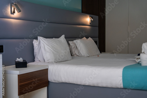 Fotografi A modern boutique hotel room in Mellieha, Malta presents a double bed with white linens and a turquoise runner, gray padded headboard, reading lights, and soft indirect lighting