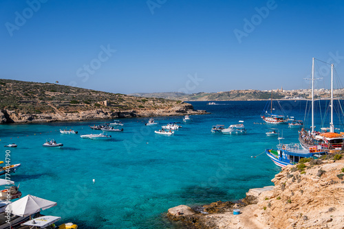 Fotografi Turquoise shallows at Blue Lagoon, Comino, Malta show boats and yachts at anchor near limestone shores, swimmers and day trippers gather under clear summer light