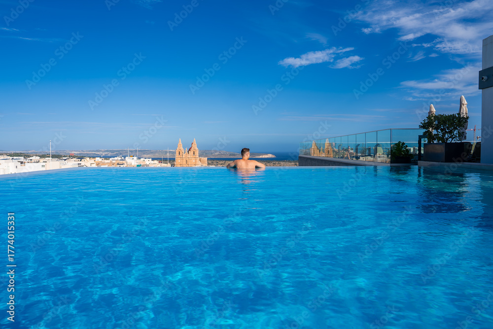 Naklejka premium A rooftop infinity pool in Mellieha, Malta, with one swimmer gazing toward the Mediterranean Sea, Gothic spires of the Sanctuary rising above town in bright midday light.