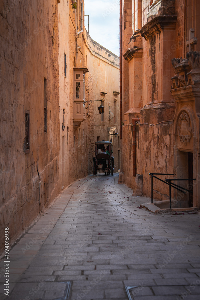Fototapeta premium A horse drawn carriage moves along cobblestones in Mdina, Malta, beside honey colored limestone, iron balconies, and arched doorways in warm afternoon light.
