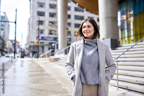 Young happy elegant brunette woman wearing coat standing on big city street. Smiling business lady walking outdoors looking away enjoying fall time in downtown on sunny day.