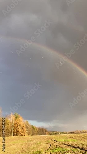 Rainbow over a golden autumn forest and field after rain. Raindrops sparkle in sunlight under dramatic gray clouds. Beautiful natural landscape, serene and atmospheric panoramic scene.