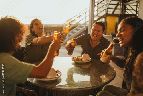 Latino friends laughing and toasting with beer bottles during golden hour at a restaurant