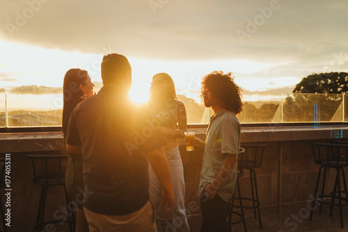 Group of young adults standing at a rooftop bar during sunset, enjoying drinks and socializing. 