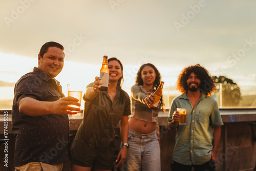 Happy group of multicultural young adults raising beers and smiling during sunset on a rooftop. Relaxed outdoor celebration, friendship and summer lifestyle concept.