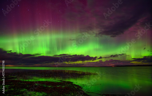 Vibrant aurora borealis in green and purple hues illuminating the night sky over a calm lake with reflections and clouds.