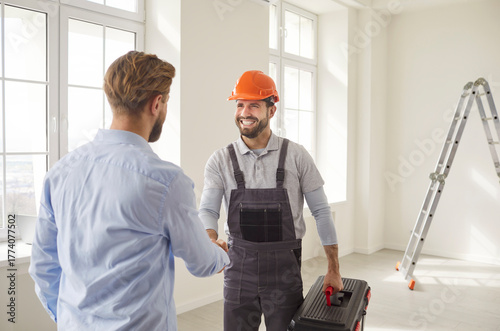 Meeting shaking hands in empty room near ladder, two men interacting, worker wearing hard hat and carrying toolbox smiling, professional repair agreement made or service trust and positive emotion 