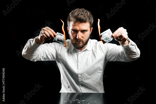 Angry man in white shirt burning money on black background, concept of financial loss, crisis, bankruptcy, bad investment, or wasting money.