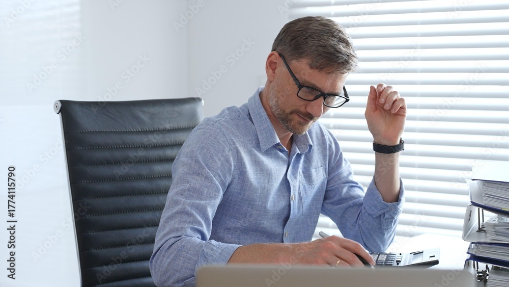 Naklejka premium Adult businessman wearing blue sleek shirt and glasses, is thinking and concentrating while working at the desk in a bright, modern office, demonstrating professionalism and efficiency. Business