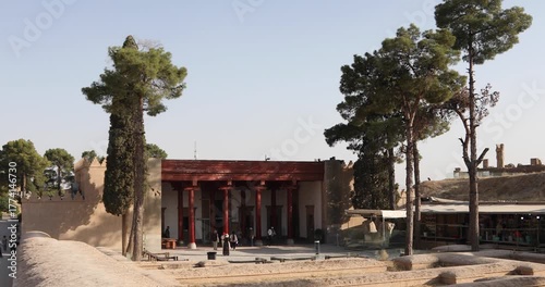 Wallpaper Mural Entrance pavilion at Persepolis, Iran, with restored red columns, tall pine trees, and a few visitors near site museum and bazaar stalls, all in warm afternoon light at the UNESCO World Heritage ruins Torontodigital.ca