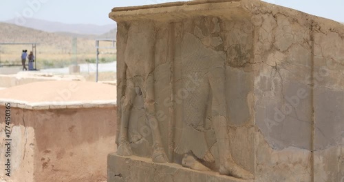 Wallpaper Mural Close-up of a weathered Achaemenid bas-relief at Pasargadae, its carved legs and robes cracked by time. Two visitors stand soft-focused near a fence in the arid landscape. Torontodigital.ca