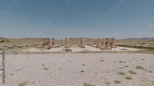 Wide view of Pasargadae’s colonnaded hall: stone columns and bases aligned across the palace floor, excavated grid visible in bright summer light with arid hills on the horizon.